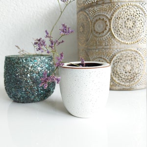 Könnte beinhalten: Zwei kleine weiße Keramik-Blumentöpfe mit gesprenkeltem Finish, einer daneben mit einem türkisfarbenen Glas-Blumentopf. Ein Zweig mit lila Blüten befindet sich im weißen Topf rechts.