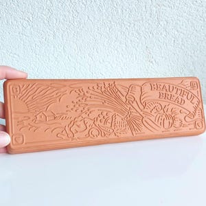 Puede incluir: Un sello de pan rectangular de color terracota con un diseño en relieve. El diseño incluye una escena rural con una casa, trigo y una pancarta que dice "BEAUTIFUL BREAD". El sello está sostenido por una mano.