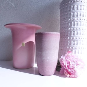 Könnte beinhalten: Drei rosafarbene Keramikvasen in verschiedenen Formen und Größen. Die Vase rechts ist strukturiert und die Vase links hat eine breite, flache Oberseite.
