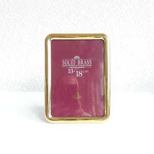 Può includere: Una cornice rettangolare in ottone massiccio color oro. La cornice ha angoli arrotondati e un retro color bordeaux. La cornice misura 13x18 cm. Il testo "SOLID BRASS" è stampato sulla cornice.