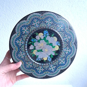 Peut inclure: Une assiette ronde décorative en étain avec un motif floral. L'assiette présente un centre noir avec un bouquet de fleurs blanches, jaunes et bleues. Le bord extérieur présente un motif bleu et sarcelle détaillé avec une bordure argentée. L'assiette est tenue par une main.