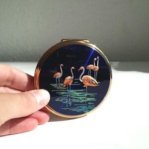 Peut inclure: Compact rond avec un bord doré et un couvercle bleu marine orné d'un motif de flamants roses. L'illustration montre plusieurs flamants roses debout dans l'eau avec des reflets verts et bleus.