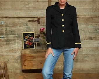 Könnte beinhalten: Eine Frau trägt einen schwarzen Blazer mit goldenen Knöpfen und eine blaue Jeans. Sie steht vor einer Holzwand mit einem kleinen gerahmten Bild und einem Schild mit der Aufschrift "We're going to be friends".