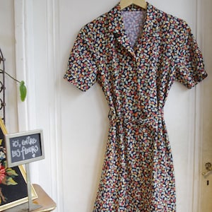 May include: A vintage style dress with a dark blue background and a colorful leaf pattern. The dress has a collared neckline, short sleeves, and a button-up front. It has a belt at the waist.