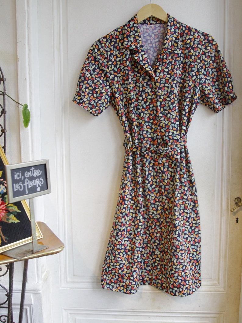 May include: A vintage style dress with a dark blue background and a colorful leaf pattern. The dress has a collared neckline, short sleeves, and a button-up front. It has a belt at the waist.