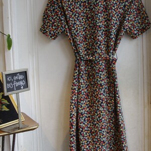 May include: A vintage-style dress with a short sleeve and a colorful leaf pattern. The dress has a tie at the waist and is made of a lightweight fabric. The dress is hanging on a white door with a sign that says "ici, entre les fleurs".