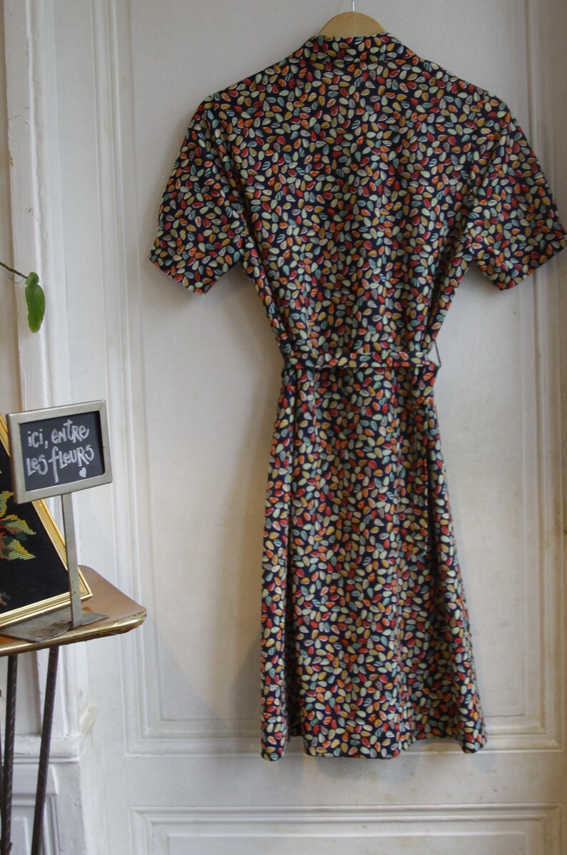 May include: A vintage-style dress with a short sleeve and a colorful leaf pattern. The dress has a tie at the waist and is made of a lightweight fabric. The dress is hanging on a white door with a sign that says "ici, entre les fleurs".