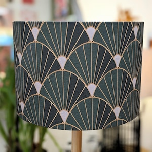 May include: A cylindrical lampshade with a dark teal and gold art deco pattern. The design features fan-like shapes in gold, with light grey accents. The lampshade is mounted on a wooden base, creating a stylish lighting fixture.