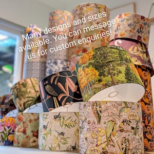 May include: A collection of lampshades with various patterns, including floral, bird, and landscape designs. The lampshades are cylindrical and stacked, showcasing a variety of colors and styles. Text on the image reads: "Many designs and sizes available. You can message us for custom enquiries!"
