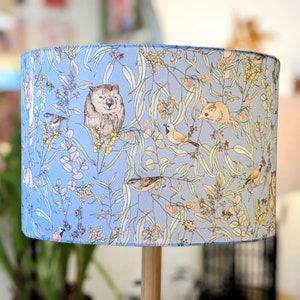Puede incluir: Una pantalla de lámpara azul claro con un patrón floral que presenta pájaros, un wombat y un opossum. El patrón está sobre un fondo azul claro.