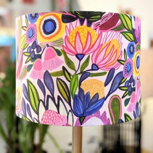 Puede incluir: Una pantalla de lámpara colorida con un patrón floral con flores rosas, amarillas, azules y verdes. La pantalla de la lámpara está hecha de tela y tiene un fondo blanco.