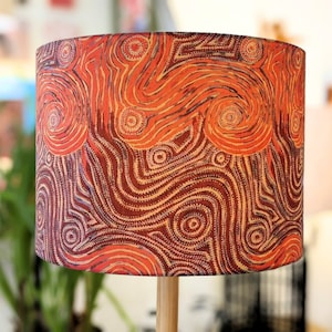 Puede incluir: Una pantalla de lámpara cilíndrica con un diseño estampado naranja y marrón. El patrón presenta líneas y puntos en espiral, que recuerdan al arte aborigen.