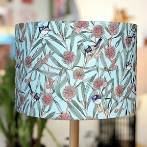 Puede incluir: Una pantalla de lámpara azul claro con un patrón floral que presenta pequeños pájaros azules y flores rosas. La pantalla de la lámpara está hecha de tela y tiene una forma cilíndrica.