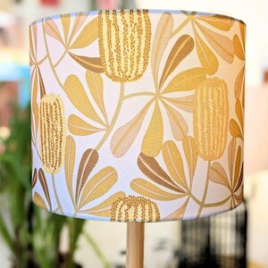 May include: A white lampshade with a floral pattern featuring yellow and brown banksia flowers. The lampshade is cylindrical and has a white background.