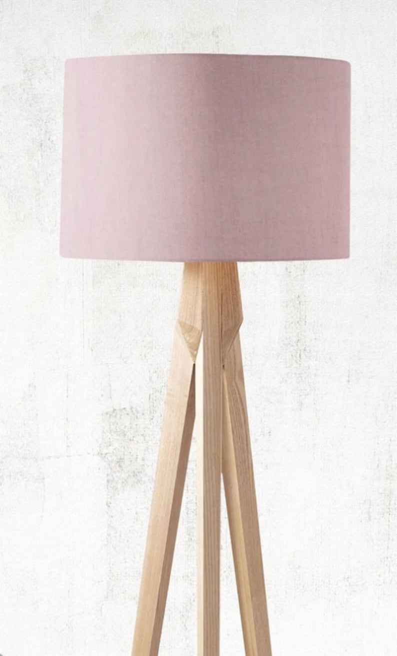 May include: A modern floor lamp with a light pink lampshade and a natural wood tripod base. The lampshade is cylindrical, and the wooden legs are angled for stability. This lamp is a stylish addition to any living space.