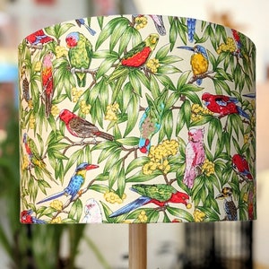 Puede incluir: Una pantalla de lámpara con un patrón tropical de pájaros y flores. La pantalla de la lámpara es de color crema con hojas verdes, flores amarillas y pájaros coloridos.