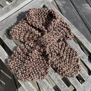 May include: A hand-knitted scarf in shades of brown, white, and orange, resting on a weathered wooden bench. The scarf features a thick, textured knit pattern, perfect for warmth and style. The scarf is a cozy accessory.
