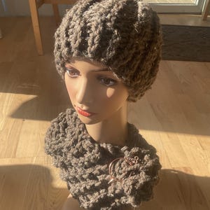 May include: A brown knitted hat and matching cowl with a decorative metal clasp. The hat and cowl are made of a thick, textured yarn.