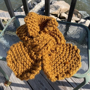 May include: A chunky knit scarf in a mustard yellow color. The scarf is made of soft yarn and has a textured, cable knit pattern.