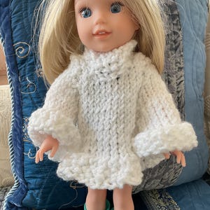 May include: A doll with blonde hair and blue eyes wearing a white knitted poncho with ruffled sleeves. The doll is also wearing turquoise rain boots. The doll is posed in front of a blue pillow.