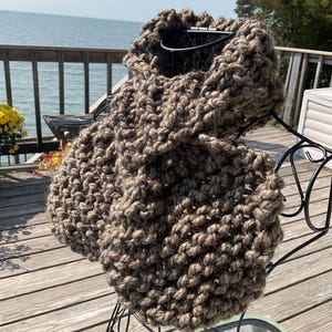 May include: A chunky, hand-knitted cowl in a neutral brown color. The thick yarn creates a textured appearance. The cowl is displayed on a decorative metal stand, with a wooden deck and water in the background.