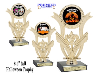 Peut inclure: Trois trophées d'Halloween dorés avec des bases noires. Chaque trophée présente une image ronde noire imprimée avec un thème d'Halloween différent. Le trophée du haut présente un crâne et des os croisés, le trophée du milieu présente un groupe d'enfants en costume et le trophée du bas présente une citrouille. Chaque trophée porte le texte "Gravure gratuite jusqu'à 2 lignes".