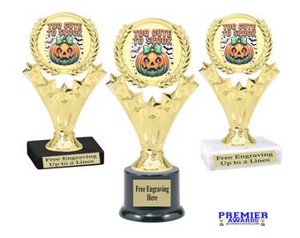 Peut inclure: Trois trophées dorés présentant un emblème circulaire avec un graphique de citrouille colorée, un nœud vert et le texte "TOO CUTE TO SPOOK" entouré de chauves-souris. Les trophées ont des accents en forme d'étoile sur des bases rectangulaires.