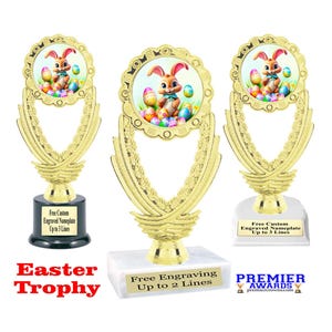 May include: Gold trophy with a wreath design and a round insert featuring an Easter bunny surrounded by colorful eggs. The trophy base is black with a white marble base. The text "Easter Trophy" is printed in red below the trophy. The text "Free Engraving Up to 2 Lines" is printed on the marble base. The text "Free Custom Engraved Nameplate Up to 3 Lines" is printed on the base of the trophy.