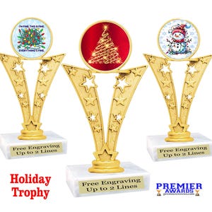 May include: Three gold-colored trophy awards with a starburst design. Each trophy has a round, white base with a black inscription that reads "Free Engraving Up to 2 Lines". The center trophy has a red circle with a gold-colored Christmas tree design. The left trophy has a blue circle with a Christmas tree design and the text "I'm fine this is fine everything's fine". The right trophy has a white circle with a snowman design. The trophies are on a white surface.