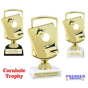 May include: Gold cornhole trophy with a white marble base. The trophy features a stylized cornhole board with a hole and a bag. The base has the text "Free Engraving Up to 2 Lines".