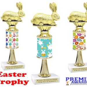 May include: Three gold Easter bunny trophies with a white base. Each trophy has a different colorful Easter egg design on the column. The text "Free Engraving Up to 2 Lines" is on each base. The text "Easter Trophy" is below the trophies.