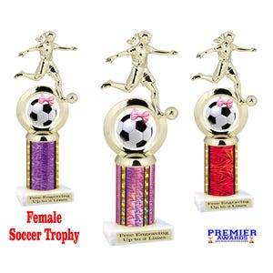 May include: Three gold soccer trophies with a female soccer player figure on top. Each trophy has a soccer ball with a pink bow, a colorful column, and a white base. The text "Female Soccer Trophy" is visible.