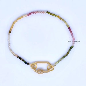 Pode incluir: Um bracelete de pedras preciosas multicoloridas com um fecho dourado. O bracelete apresenta um degradê de cores, começando com amarelo e passando para rosa, depois verde. O fecho é um oval dourado com uma fila de pedras transparentes.
