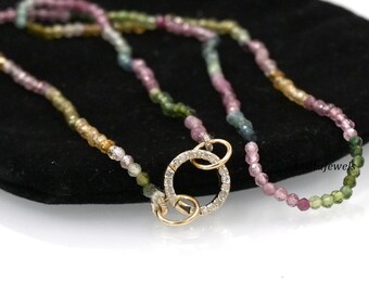 May include: A multi-colored gemstone beaded necklace with a gold clasp featuring a circle of small diamonds. The necklace is on a black velvet surface.