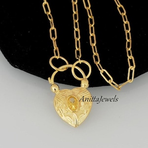 May include: A gold heart-shaped locket necklace with a small yellow gemstone in the center. The necklace has a chain link design and a clasp closure. The locket is engraved with floral details. The text "AnittaJewels" is visible in the image.
