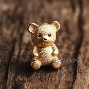 May include: A gold-colored teddy bear figurine with a textured surface. The bear has small, round ears, a smiling face, and is sitting with its arms at its sides. The background is a blurred wooden surface.