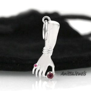 Op de afbeelding: Zilveren bedel van een hand die een roze edelsteen vasthoudt. De hand is gedetailleerd met een armband en een ring met een roze edelsteen.