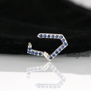 Puede incluir: Un pendiente de aro de plata con forma geométrica, adornado con pequeñas piedras azules. El aro es abierto y está diseñado para ajustarse cómodamente a la oreja.