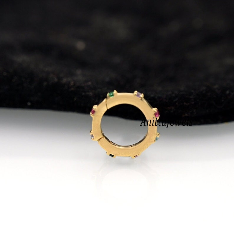 Op de afbeelding: Gouden creolen oorring met een scharnier sluiting en kleurrijke edelstenen rond de rand. De oorring is op een zwarte achtergrond.