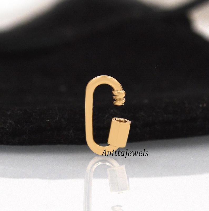 May include: Gold-toned metal clasp with a rounded oval shape. The clasp has a spring-loaded mechanism and a small, cylindrical piece on one side.