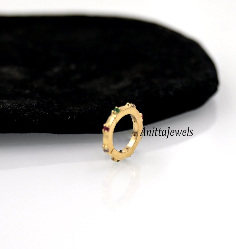 Op de afbeelding: Een gouden ring met een rij kleurrijke edelstenen. De ring ligt op een zwart fluwelen oppervlak. De tekst "Anittajewels" is zichtbaar in de afbeelding.