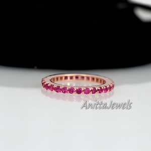 Op de afbeelding: Een roségouden bandring met een rij kleine, ronde, roze edelstenen. De ring ligt op een wit oppervlak met een zwarte achtergrond.