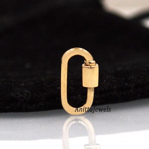 May include: Gold-toned metal lobster clasp charm. The clasp is oval-shaped with a small, cylindrical closure. The charm is on a black background.