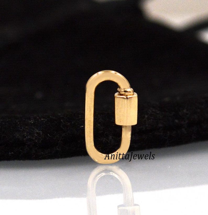 May include: Gold-toned metal lobster clasp charm. The clasp is oval-shaped with a small, cylindrical closure. The charm is on a black background.