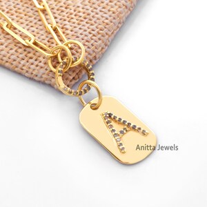 Könnte beinhalten: Goldkette mit einem kleinen, rechteckigen Anhänger, der den Buchstaben 'A' mit kleinen Diamanten umrandet zeigt. Der Anhänger ist mit einem kleinen, runden Ring an der Kette befestigt. Der Text 'Anitta Jewels' ist in der unteren rechten Ecke des Bildes sichtbar.