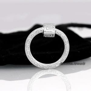 May include: A silver-colored ring with a Greek key pattern around the band and a rectangular centerpiece. The ring is displayed on a reflective surface with a black background.