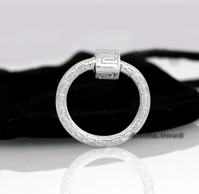 May include: A silver-colored ring with a Greek key pattern around the band and a rectangular centerpiece. The ring is displayed on a reflective surface with a black background.