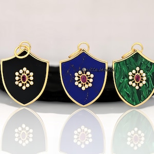 May include: Three gold shield pendants with a gemstone center surrounded by a circle of white stones. The shields are black, blue, and green.