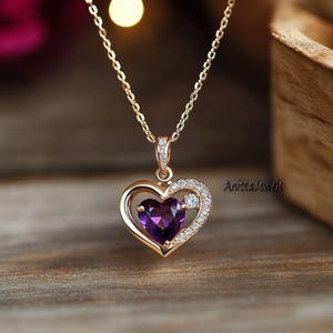 Puede incluir: Un collar de oro con un colgante en forma de corazón, adornado con una piedra preciosa púrpura en el centro y pequeñas piedras preciosas transparentes alrededor del borde. El collar está en una cadena de oro.