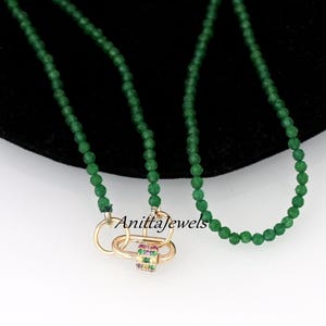 Puede incluir: Un collar de cuentas de piedras preciosas verdes con un cierre dorado que presenta un acento de piedra multicolor. El collar está sobre un fondo negro. AnittaJewels está escrito en el cierre.